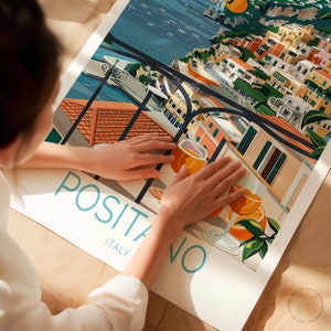 May include: A colorful illustration of Positano, Italy, with a view of the town from a balcony. The illustration features a glass of orange juice and a lemon tree. The text "Positano, Italy" is written in a blue font.