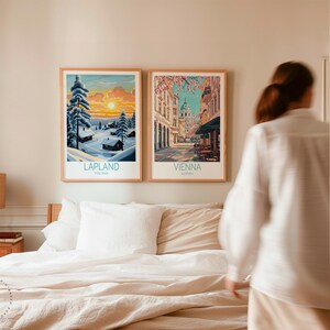 May include: Two framed travel art prints. One shows a winter scene in Lapland, Finland, with snow-covered trees and a sunset. The other depicts a street scene in Vienna, Austria, with buildings and cherry blossoms. Both prints have wooden frames.