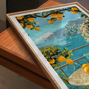 May include: A framed print of a colorful illustration depicting a view of the Italian city of Sorrento. The illustration features a balcony overlooking the Mediterranean Sea with a glass of lemonade and a plate of lemons on a table. The text "SORRENTO" is written in white letters on a blue background.