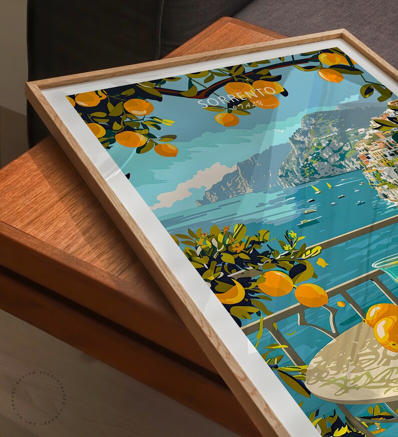 May include: A framed print of a colorful illustration depicting a view of the Italian city of Sorrento. The illustration features a balcony overlooking the Mediterranean Sea with a glass of lemonade and a plate of lemons on a table. The text "SORRENTO" is written in white letters on a blue background.