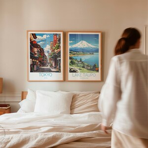 May include: Two framed travel posters on a wall above a bed with white linen. The poster on the left shows a street scene in Tokyo, Japan. The poster on the right shows a mountain and lake scene in Lake Taupo, New Zealand.