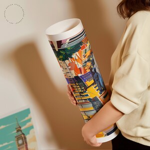 May include: A rolled up poster featuring a colorful illustration of a city scene with buildings, streets, and people. The poster is being held by a person's hand.