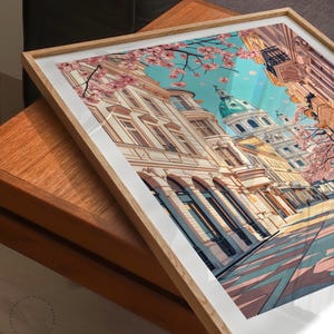 May include: A framed art print depicting a stylized cityscape with buildings and cherry blossoms. The artwork has a light wood frame and a white mount. The buildings are beige and white, with a blue sky and pink blossoms.