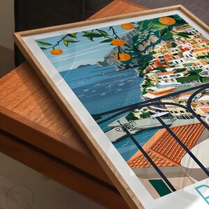 May include: A framed illustration of a colorful cityscape with a view of the ocean. The city is built on a cliffside with a lush green tree with orange fruit hanging over the edge. The ocean is blue with white boats and the sky is a light blue with white clouds.