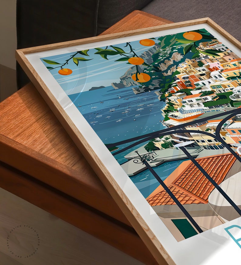 May include: A framed illustration of a colorful cityscape with a view of the ocean. The city is built on a cliffside with a lush green tree with orange fruit hanging over the edge. The ocean is blue with white boats and the sky is a light blue with white clouds.
