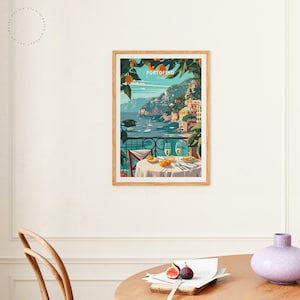 K&ouml;nnte beinhalten: Ein gerahmtes Poster mit einer farbenfrohen Illustration einer K&uuml;stenstadt mit Geb&auml;uden auf einer Klippe mit Blick auf ein blaues Meer. Das Poster tr&auml;gt den Text "PORTOFINO" in einem wei&szlig;en Kasten mit rotem Rand. Das Poster h&auml;ngt an einer wei&szlig;en Wand.