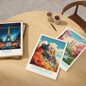 May include: Three travel art prints on a wooden table. One framed print shows the Eiffel Tower in Paris, France. The other two prints depict landscapes of Edinburgh, Scotland, and a park scene. The prints have a colourful, illustrative style.