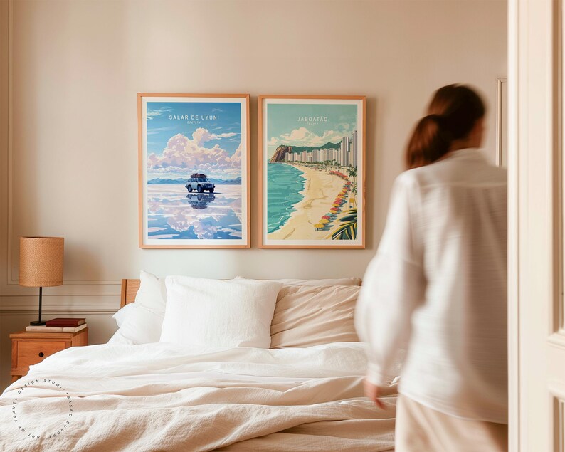 May include: Two framed travel posters hanging on a white wall above a bed with white and beige bedding. The poster on the left shows a car driving on a salt flat with the text "Salar de Uyuni, Bolivia". The poster on the right shows a beach scene with the text "Jaragu&aacute;, Brazil".