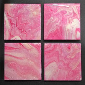 Puede incluir: Cuatro posavasos cuadrados de cerámica con un diseño abstracto en espiral rosa y blanco. Los posavasos tienen un acabado brillante y un efecto marmolado, con sutiles acentos de purpurina. La paleta de colores incluye tonos de rosa y blanco.