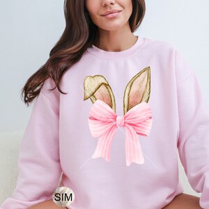 May include: Pink sweatshirt with a graphic of bunny ears with a pink bow. The bow has white stripes.