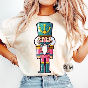 May include: A white t-shirt featuring a cartoon illustration of a nutcracker. The nutcracker is wearing a green hat with gold accents, a pink jacket, and blue trousers. The t-shirt has the word "SIM" printed on the bottom right corner.
