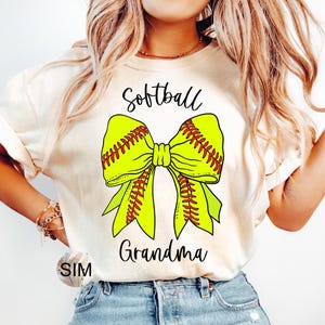 Könnte beinhalten: Ein weißes T-Shirt mit dem Text "Softball Grandma" und einer gelben und roten Softball-Schleife.