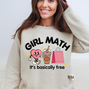 May include: A beige sweatshirt with the text "Girl Math it's basically free" in black. The graphic features a pink heart with a face, a brown iced coffee with a straw, and a pink shopping bag.