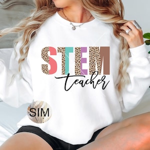 May include: White sweatshirt with the text "STEM teacher" in colorful letters with a leopard print design. The letters are in pink, teal, purple, and brown.