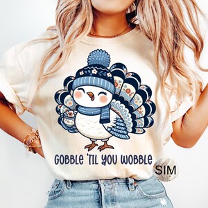 May include: Cream-coloured t-shirt with a cartoon turkey wearing a blue hat and scarf, holding a cup. The turkey has blue and white patterned feathers. The navy blue text "GOBBLE 'TIL YOU WOBBLE" is printed below the turkey.