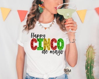 Puede incluir: Camiseta blanca con el texto "Happy Cinco de Mayo" en letras brillantes de colores. La persona sostiene una copa de margarita. Decoraciones festivas: un pendiente de cactus y una guirnalda.