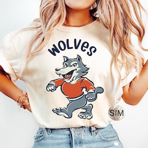 K&ouml;nnte beinhalten: Ein wei&szlig;es T-Shirt mit einem Cartoon-Wolf, der ein rotes Shirt tr&auml;gt und das Wort "WOLVES" dar&uuml;ber gedruckt ist.