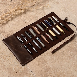 May include: A brown leather pen roll with 12 slots for pens and pencils. The roll is unrolled and the slots are filled with various writing instruments.