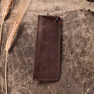 May include: A brown leather glasses case with a stitched edge. The case is lying on a marble surface.