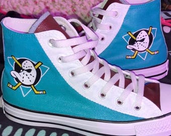 May include: Pair of high-top sneakers in teal, white, and brown, featuring the Anaheim Ducks logo. The shoes have white laces and a white rubber sole. The design includes the team's mascot on the side of each shoe.