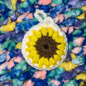 May include: A crocheted sunflower with a white center, yellow petals, and a brown center. The sunflower is on a colorful fuzzy background.