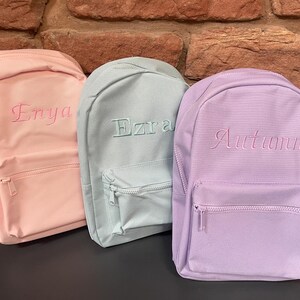 May include: Three pastel-coloured backpacks. The pink backpack has "Enya" embroidered in pink. The light blue backpack has "Ezra" embroidered in a matching colour. The purple backpack has "Autumnn" embroidered in pink. Each backpack has a front zipped pocket.