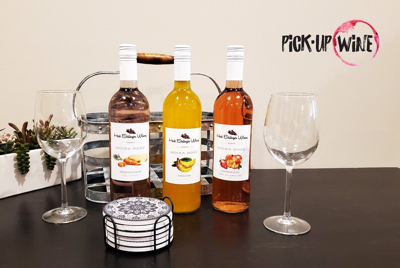 May include: Three bottles of Moira Rose wine, a pink, a yellow, and a peach color, with labels featuring fruit illustrations. The bottles are on a dark wooden table with two wine glasses and a set of coasters.