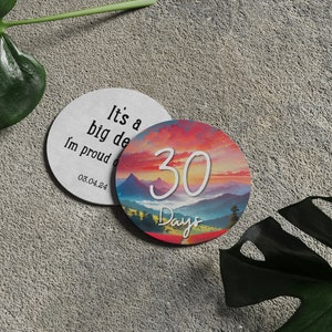 May include: Two round white coasters with a colorful mountain landscape and a sunset sky. One coaster says "It's a big deal, I'm proud of you" and "03.04.24". The other coaster says "30 Days" in white text.