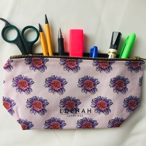 Puede incluir: Un estuche de lápices rosa con cremallera, con un patrón floral y el texto "Edenah Cambridge" impreso en la parte delantera. El estuche está lleno de varios útiles de escritura, incluidos bolígrafos, lápices y un resaltador.