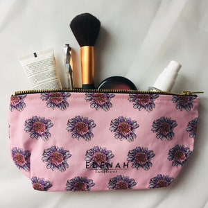 Puede incluir: Bolsa de maquillaje rosa con estampado floral y cierre de cremallera. La bolsa tiene un logotipo negro que dice "EDENAH CAMBRIDGE". La bolsa está llena de pinceles de maquillaje, un tubo de loción y una botella de spray.