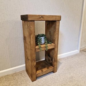 May include: A two-tiered wooden plant stand with a natural finish. The stand has a rectangular top and base, connected by vertical supports. A small potted plant sits on the middle shelf. The stand is designed for indoor use. The dimensions are approximately 60cm high.