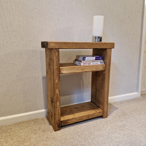 May include: A wooden side table with a warm, brown finish. The table has two shelves and a rectangular top. A small lamp and two books are on the top shelf. The table is approximately 61 cm tall.