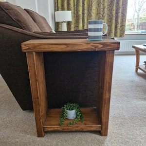 May include: A rustic wooden side table with a single shelf. The table is stained a dark brown colour and has a natural wood grain. A small potted plant sits on the shelf, and a mug with a striped pattern sits on the top shelf.