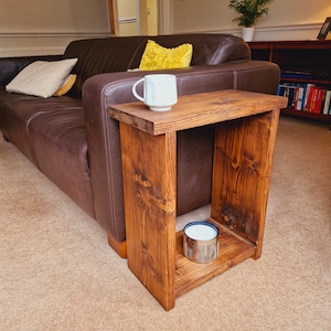 May include: A rustic wooden side table with a shelf, featuring a white mug and a candle on the shelf. The table is next to a brown leather sofa with a yellow pillow.