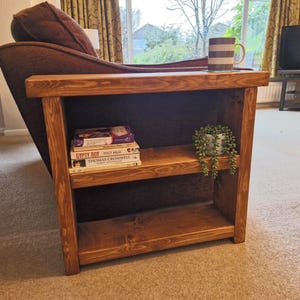 May include: A wooden side table with two shelves, stained a warm brown. It holds books, a small potted plant, and a mug. The table is next to a brown sofa. The table's dimensions are approximately 60cm in height, 50cm in width, and 30cm in depth.