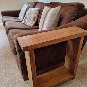 May include: A wooden side table with a shelf, stained in a dark brown colour. The table is next to a brown sofa with cushions.