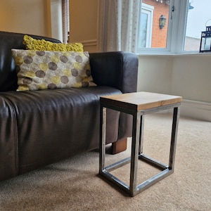 May include: A small, square side table with a wooden top and a metal frame. The table is approximately 46 cm tall and 41 cm wide. The table is in a living room setting, next to a brown leather sofa.