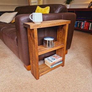 May include: A wooden side table with three shelves. The top shelf has a white mug and a candle. The middle shelf has a candle. The bottom shelf has three books. The table is next to a brown leather sofa.