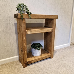 May include: A wooden side table with two shelves, made from light brown, unfinished wood. The table has a simple design and is suitable for indoor use. Two small potted plants are placed on the top and bottom shelves. The table's dimensions are approximately 60cm high.
