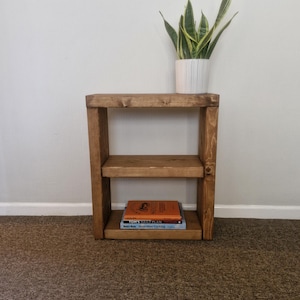 May include: A two-tiered wooden side table with a natural finish. The table has a rectangular shape with two shelves. A white potted plant with green and yellow leaves sits on top. Two books are on the lower shelf.