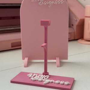 May include: A pink display stand featuring the text "angie designsss" in white script. The stand has a rectangular base and an arched back, with a central support structure. The design is a light, monochromatic pink.