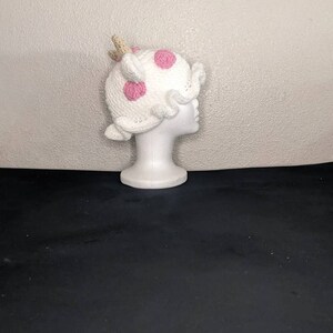 May include: White crocheted hat with pink polka dots and a brown horn on top. The hat is on a white mannequin head.