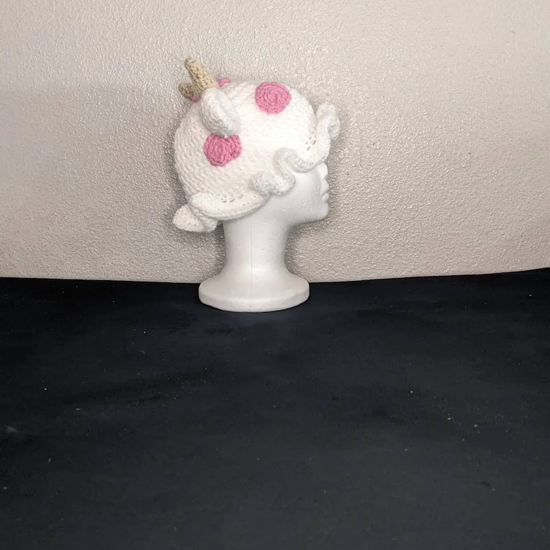 May include: White crocheted hat with pink polka dots and a brown horn on top. The hat is on a white mannequin head.
