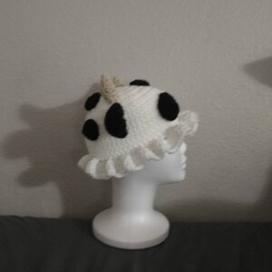 May include: A white crocheted hat with black spots and a ruffled edge. The hat is on a white mannequin head.