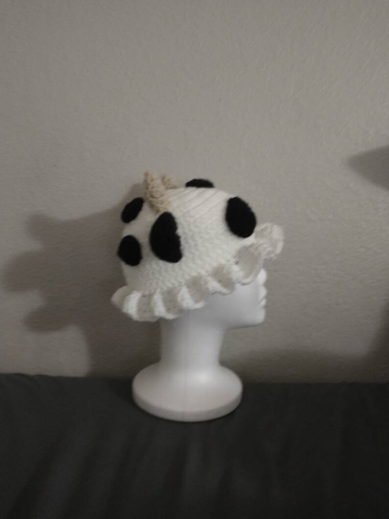 May include: A white crocheted hat with black spots and a ruffled edge. The hat is on a white mannequin head.