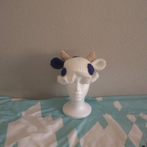 May include: A white crocheted cow hat with blue spots and brown horns. The hat is on a white mannequin head.