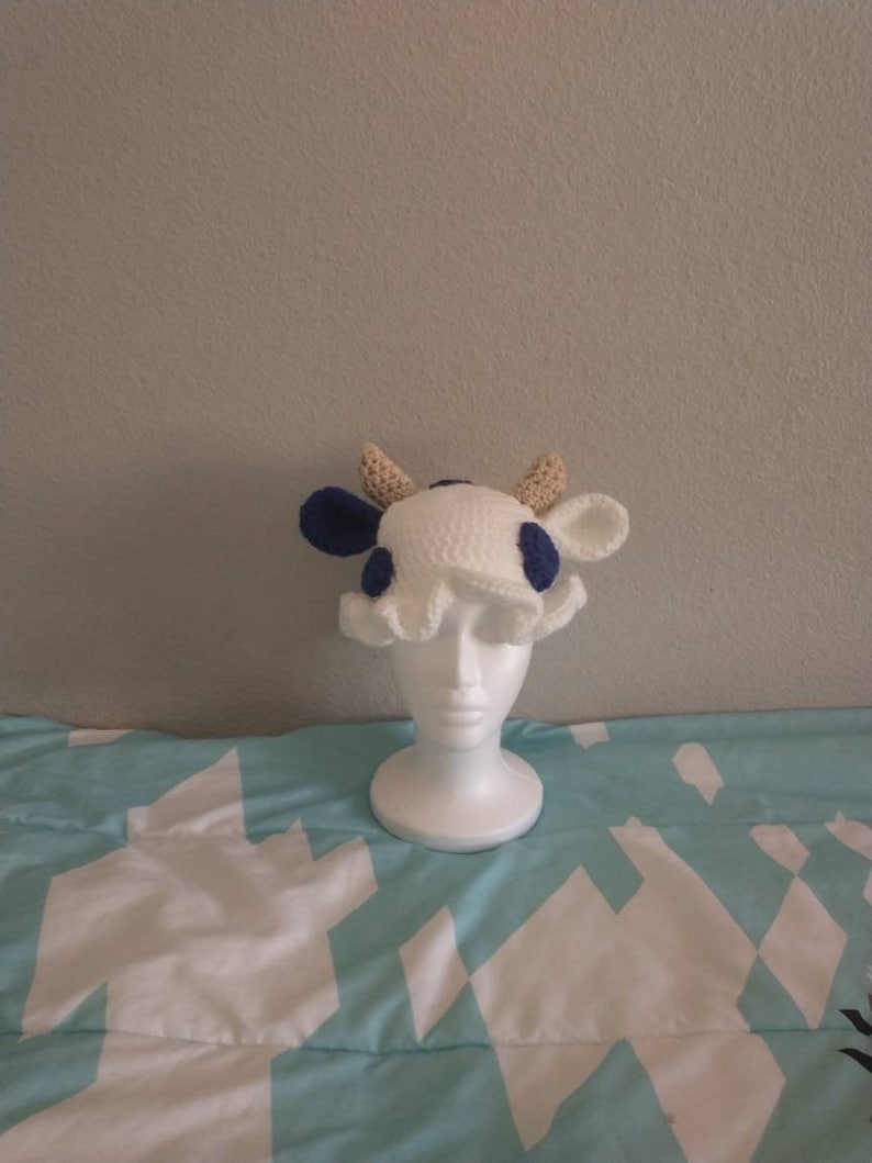 May include: A white crocheted cow hat with blue spots and brown horns. The hat is on a white mannequin head.