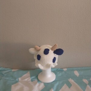 May include: White crocheted cow hat with blue spots and brown ears. The hat is displayed on a white mannequin head.