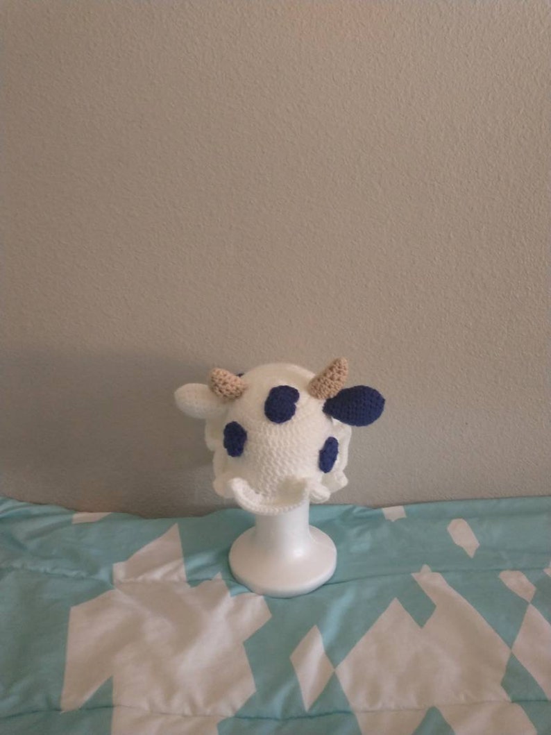 May include: White crocheted cow hat with blue spots and brown ears. The hat is displayed on a white mannequin head.