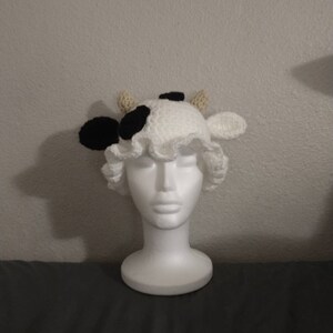 May include: White crocheted cow hat with black spots and brown horns. The hat is on a white mannequin head.